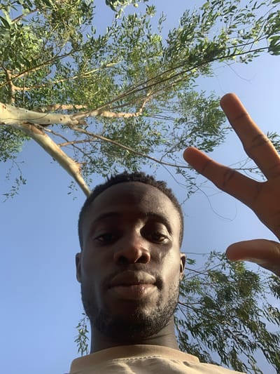 A low-angle selfie taken outdoors in daylight, featuring a dark-skinned man with short curly hair and a beard. The man is positioned in the lower right of the frame, with his face partially visible. His right hand is held above his head, fingers extended in a gesture. Behind him, a tree with slender branches and light green leaves fills the left side of the image. The background is a clear blue sky. The location is Gbeleko, Nigeria.