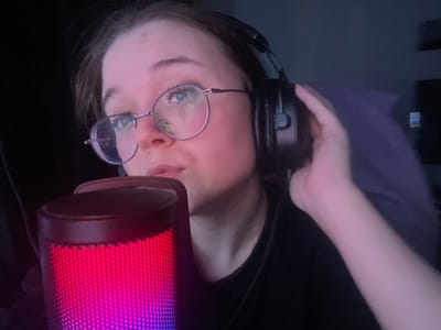 A young adult female is depicted in a close-up, indoors, under low light conditions. She has light skin, dark hair, and wears round, wire-frame glasses with light-colored lenses. Her eyes are directed upwards and to the viewer's left. She wears black over-ear headphones, with her left hand holding the left earcup against her ear. Her mouth is slightly open. In the lower-left foreground, a cylindrical microphone with a mesh grille displays a gradient of red, pink, and purple illumination. The individual is wearing a dark, presumably black, top. The background is dark and indistinct, with a dark blue/purple fabric visible behind her right shoulder. The specific location within Pushchino, Russia, cannot be determined from the visual information provided.