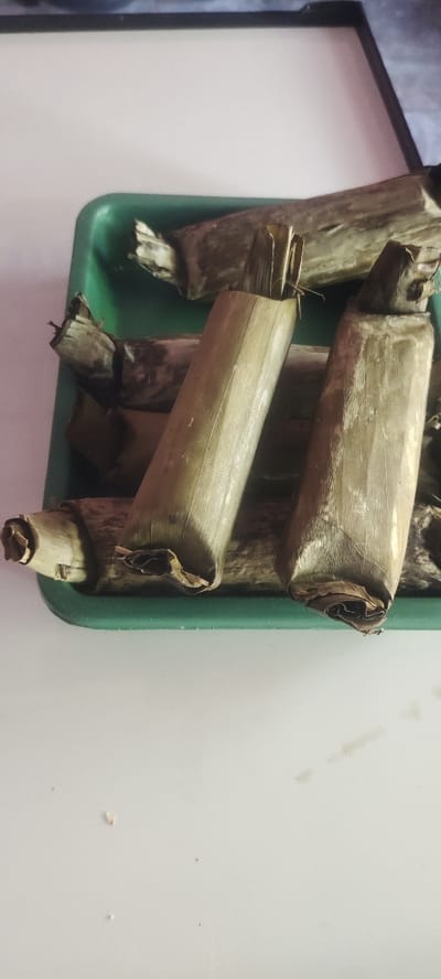The image displays an overhead, slightly angled view of approximately ten Indonesian food items, consistent with lemper, contained within a dark green, rectangular plastic tray with raised edges and rounded corners. Each food item is a cylindrical parcel wrapped in dried, light brown to olive-green leaf material, most likely banana leaves, with the ends folded and secured. The items are arranged haphazardly within the tray. The tray rests on a light-colored, possibly white, flat surface. The upper background features a blurred dark object against a lighter, undefined vertical surface. The scene is illuminated by diffuse ambient light. This specific scene is located in the city of Mojokerto, Indonesia.