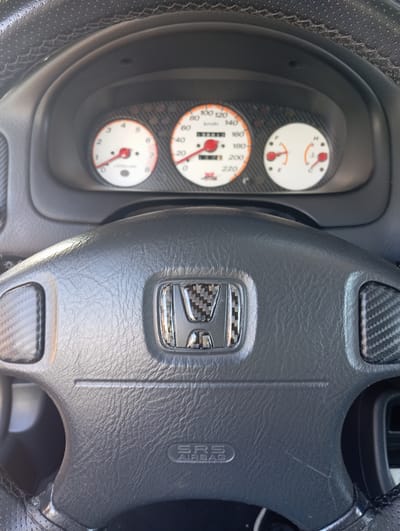 This is a close-up, high-angle shot of a car's interior, focusing on the steering wheel and dashboard. The steering wheel is made of dark leather with textured accents, likely carbon fiber, on the sides and center, where a Honda emblem is prominently displayed. Below the emblem, "SRS AIRBAG" is embossed on the steering wheel. The dashboard features three circular gauges, one for speed (reading approximately 100 km/h), one for RPMs, and a smaller gauge that appears to indicate fuel level. The background is blurred, but suggests the interior of a vehicle. The image is crisp and well-lit, with no visible people or activity outside the car. The location in the city of Arma, Italy, is not discernible from the image itself.