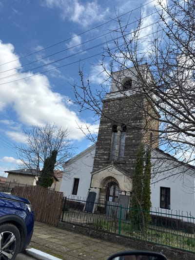 The image shows the St. Nicholas Church in Kameno Pole, Bulgaria. The church has a white facade, a gray stone bell tower, and an arched entrance covered with a metal fence. A tree with bare branches is in the foreground, obscuring parts of the church. Electrical wires are visible against the blue sky with white clouds. A blue car is partially visible in the lower right corner, parked on a paved street. A wooden fence is visible near the car. The overall scene appears to be taken from a low angle, looking up at the church.