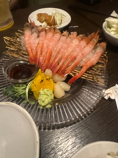 The image shows a close-up of a platter of raw shrimp served as sashimi. The platter is clear plastic and sits on a dark wooden table. The shrimp are arranged neatly, with a small bowl of dipping sauce, pickled vegetables, wasabi, sprouts, and shredded daikon radish also present. Other plates are visible on the table. The location is Gimpo-si, South Korea.