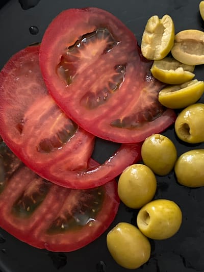 The image shows a close-up, top-down view of sliced tomatoes and green olives arranged on a black, reflective surface. Two large, red tomato slices are prominently featured, with their seedy interiors and glistening moisture visible. Scattered around the tomatoes are several whole and halved green olives. The surface appears to be a plate or tray. There is no indication of the city of Pushkino, Russia, in the image. The lighting is direct, creating highlights and some shadows. The scene is still, with no active elements.
