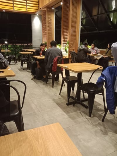 The scene takes place inside a cafe, located in Kecamatan Ngaliyan, Indonesia (latitude: -7.013, longitude: 110.338). The cafe features a tiled floor, wooden tables, black metal chairs, and wooden walls with slatted panels. It appears to be a well-lit establishment with large windows offering a view of the outside. Four individuals are seated at separate tables, engaged in conversation. One individual is holding a drink with a straw. A blue jacket is draped over the back of a chair. The tables are arranged in a staggered formation, with a partially obscured table in the foreground.  The cafe is a place of social interaction and refreshment.