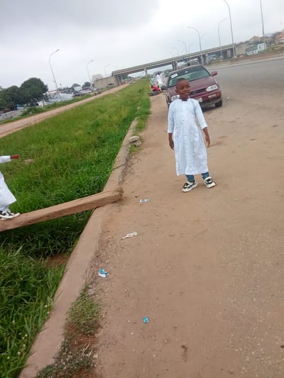 A young boy, dressed in a white kaftan and jeans, stands on a dirt path beside a grassy embankment. He is looking towards the camera with a slight smile. In the background, a maroon car is parked on the side of a road. Further in the distance, an elevated highway passes overhead, with cars visible on it. The sky is overcast, suggesting a cloudy day. The scene appears to be in a semi-urban or rural area in Kagini, Nigeria, given the mix of road infrastructure and natural elements. There is a wooden plank laid across a ditch, and the boy's right foot is near it. The overall atmosphere is casual and everyday.