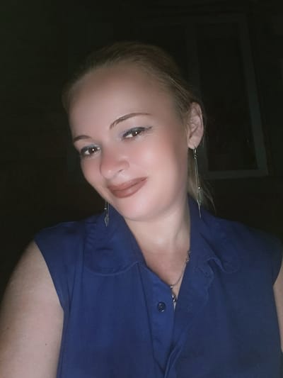 The image shows a close-up, head-and-shoulders shot of a fair-skinned woman with blonde hair pulled back. She is wearing a dark blue sleeveless collared shirt. She has on eye makeup, including what appears to be blue eyeshadow, and lipstick. She is wearing long, dangling earrings. The background is dark and out of focus, with what appears to be a window frame visible on the right side of the image. The lighting suggests the photo was taken indoors at night. There is no indication of the location being in Chernihiv, Ukraine.