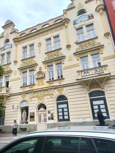 An exterior, eye-level, medium shot captures the ornate facade of a building in Pardubice, Czechia, likely a residential or commercial property, with a light yellow exterior and detailed yellow and green decorative elements. The architecture features a balcony, arched windows, and what appears to be a mural or fresco above some of the windows. At the ground level, there is a shop with the name "PARADIS Boutique" and a mannequin displayed in the window. A woman in a grey jacket and dark pants is walking past the shop. The foreground shows the top of a silver car, partially obscuring the lower portion of the building. The sky is overcast.