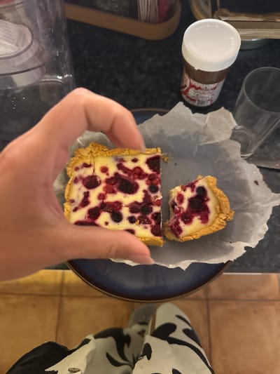 A hand holds a slice of berry tart above a blue plate containing the remainder of the tart. The tart has a shortbread-like crust and a creamy filling studded with red and dark purple berries. The plate is lined with parchment paper. In the background, there is a jar of Nutella, a glass, and part of a patterned, likely animal print, garment. The setting appears to be indoors on a countertop. There's nothing to indicate that this is in Miljevci, Montenegro.