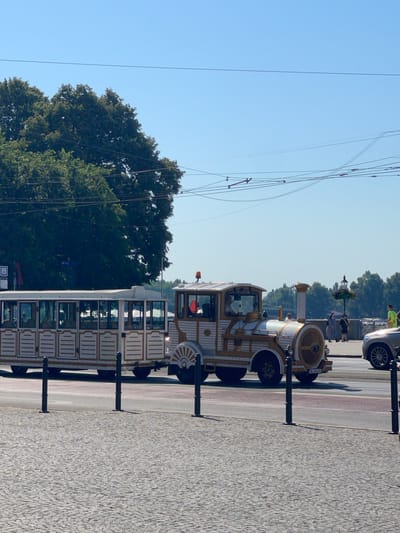 The image captures a sunny day in Bratislava, Slovakia, specifically along the Danube River promenade. In the foreground, a white and gold tourist train, designed to resemble a vintage locomotive and carriages, is parked on the cobblestone pathway. The train has large windows, and several people appear to be seated inside. The locomotive section features a distinctive round grill and an orange light on its roof.

Behind the train, the paved promenade is visible, with several black bollards separating it from the road. Pedestrians can be seen walking along the promenade, including a person in a nun's habit and a person wearing a bright yellow shirt. A modern silver car is parked on the road adjacent to the promenade.

In the background, lush green trees form a dense canopy, with the blue sky stretching above. Overhead, power lines are strung across the sky, indicating the presence of a tram or trolleybus system. The overall scene suggests a pleasant tourist activity taking place on a bright, clear day in the Slovakian capital.