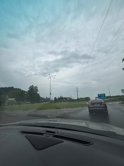 The image depicts a view from inside a vehicle looking out at a wet roadway and surrounding environment under an overcast sky. The foreground shows the dashboard of the car. In the midground, a dark-colored sedan with license plate PT-059-TT is driving on the right side of a multi-lane intersection or roundabout. The road appears wet, suggesting recent rain. Various traffic signs are visible, including directional signs in Georgian. The background features green trees and rolling hills, indicative of a rural or semi-rural setting. A tall street lamp with multiple lights is present on the left side of the road. Power lines are visible stretching across the sky. The location is identified as ნიგოითის თემი, Georgia.