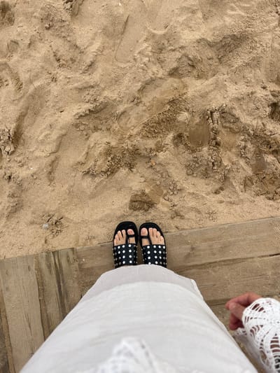A high-angle, downward view depicts the lower portion of a person's body at a beach in Santander, Spain. The individual is wearing a light-colored, possibly white, garment, with a segment of white lace visible on the lower right side, suggestive of a sleeve or cuff. Two bare feet are present, each inserted into a black slide-style sandal. Both sandals feature a broad top strap embellished with multiple small, round, metallic studs. The left sandal incorporates a toe loop, while the right sandal features a toe post. The sandals are positioned on a light brown wooden boardwalk composed of horizontally oriented planks. Beyond the boardwalk, occupying the majority of the upper frame, is an expanse of light brown beach sand exhibiting irregular textures, ripples, and depressions.
