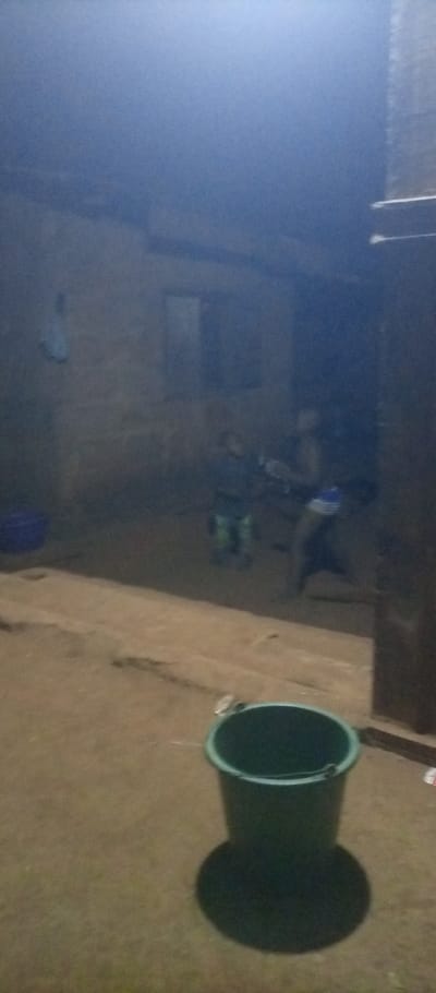 A dimly lit outdoor scene features a dark green bucket in the foreground, casting a distinct shadow on the ground. In the background, two figures are vaguely visible near a building with a window. A blue container sits to the left of the structure. The overall image has a grainy quality and poor visibility, suggesting low light conditions or a low-resolution photograph.
