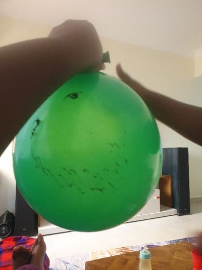 The image depicts an indoor scene in Mumbuni North ward, Kenya. A large, inflated green balloon is centrally positioned, held at the top by two dark-skinned hands. The left hand grasps the balloon's neck, while the right hand rests on its upper-right surface. The balloon has several dark, irregular markings and a small black drawn 'eye' shape near the top. In the bottom-left corner, a pair of bare, dark-skinned feet rests on a red and blue patterned fabric, likely a blanket or throw. The background reveals a domestic setting: a light-colored tiled floor, light-beige walls, and various objects. On the floor to the left, a black portable speaker is visible next to a larger black floor-standing speaker. Another matching large black speaker stands further right in the background. A wooden coffee table with a decorative top is in the foreground on the right, featuring a baby bottle with a light-colored liquid and a green nipple, and a partial view of a red container.