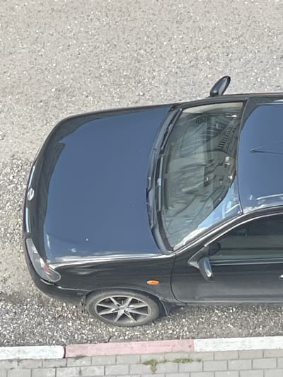 The image shows a dark-colored car parked on a gravelly surface next to a sidewalk made of light-colored bricks. The car is viewed from a high angle, looking down. The front of the car is visible, including the hood, windshield, and part of the driver's side mirror. The windshield reflects a building with multiple windows. The side of the car, including a wheel and an amber turn signal, is also visible. The scene appears to be outdoors, with the lighting suggesting it is daytime. There is no visible text in the image. The overall impression is of a static, everyday scene of a parked vehicle.