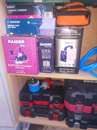 The image shows a shelf with various tools and toolboxes. On the top shelf, there are two boxes of power tools. The first box on the left is for a "Parkside 2-Speed Drill / Screwdriver" and the second box on the right contains a "Parkside Digital Hot Air Gun". In the middle shelf, on the left, is a pink box labeled "RAIDER Pro Electric Planer RDP-EP15". Next to it is a blue box for "RAIDER Power Tools". To the right of that is a box for a "WERSTIL Inverter MMA Welding Machine MMA350" with a blue welding cable coiled beside it. The bottom shelf contains two black and red toolboxes, one smaller one on the left and a larger one on the right. A red and black impact driver and a green and blue grinding disc are placed on top of the smaller toolbox. The overall environment appears to be a storage area or workshop. There is no indication of the location being in the city of Karnobat, Bulgaria.
