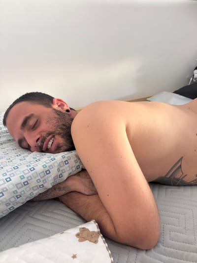 A man lies supine on a bed with a blue-and-white patterned pillow under his head. His arms are crossed, partially obscuring his face. He has a beard, dark hair, a nose ring, and tattoos on his chest and arms. He appears to be asleep or resting, with his eyes closed and mouth slightly open. The bed is covered with a grey textured blanket and a white blanket with a teddy bear design. The background consists of a plain white wall. The location is unknown and not specified.
