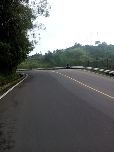A winding asphalt road with a solid yellow line running down the center curves through a hilly landscape. A metal guardrail lines the road's edge on one side. Trees and foliage border the other side of the road. A lone motorcyclist sits on the side of the road. Towers are visible in the distance atop a grassy hill. The sky is overcast.