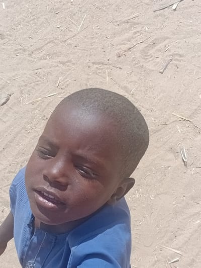 A young child, located in Ayama, Nigeria, is depicted from a high-angle, looking upwards towards the viewer. The child presents with dark skin, short dark hair, and is wearing a blue short-sleeved garment with vertical textured stitching on the front and a faint light-colored design near the neckline. The child's eyes are partially closed, appearing to squint, and the mouth is slightly open, revealing some teeth. The background consists of light tan-colored sand, marked with scattered fragments of dry organic material, such as twigs and straw, along with faint surface impressions. The scene is brightly illuminated, suggesting direct overhead sunlight.