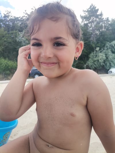 This is a close-up, waist-up outdoor photograph of a young girl on a beach. The girl has dark, curly hair, dark eyes, and fair skin. She is smiling broadly and touching her right ear with her left hand. She is wearing a small, gold hoop earring with a pink heart-shaped gem in her right ear. Her shoulders and upper chest are lightly dusted with sand.

The background consists of a sandy beach, blurred green trees, and a cloudy sky. In the far left of the image, a portion of a blue plastic toy bucket with a cartoon octopus design is visible. In the background, a dark-colored car and a white car are partially visible.

The scene appears to be set on a beach during the daytime, with overcast or partly cloudy weather. The girl's attire and the sandy environment suggest she is at the beach for leisure, possibly swimming or playing in the sand. Her expression conveys happiness and playfulness.

There is no visible text in the image. The location context of Kranevo, Bulgaria, places the scene in a coastal resort town.