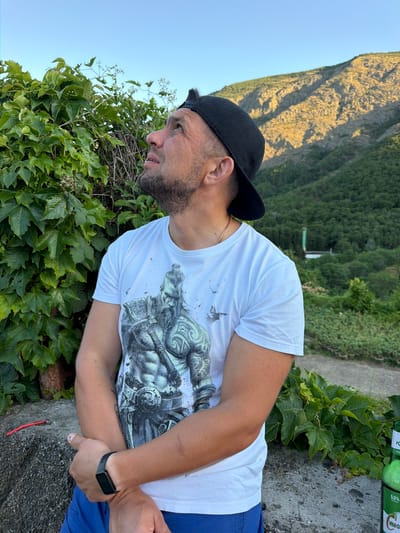 This media file shows a man wearing a white t-shirt with a graphic print, a black baseball cap, and blue shorts. He has a beard and is wearing a black smartwatch on his left wrist. The man is looking off to the side, with a neutral expression. He appears to be in an outdoor setting with a rocky embankment and lush green foliage. In the background, a steep, sunlit mountain covered in vegetation rises against a clear sky. The scene suggests it is daytime, likely late afternoon due to the angle of the sunlight illuminating the mountain. Two green bottles with visible text are placed on the embankment behind the man. Part of the text on one bottle appears to read "КИЇВСЬКЕ" and on another "5.0%". The overall impression is a casual moment outdoors, possibly during a trip or a break in a scenic location.