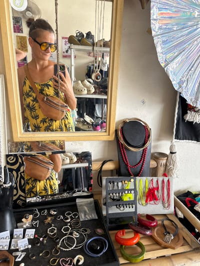 The media file captures a cluttered, eclectic display of accessories, possibly in a market stall or personal collection, with a young woman reflected in a mirror. The setting appears to be indoors, with a textured, light-colored wall as the backdrop. Prominently featured are various jewelry items, including necklaces with red beads, bracelets in assorted colors like red, green, and orange, and an assortment of rings and earrings displayed in black trays. There are also bundles of red and yellow threads and what look like handmade crafts or decorations, including a large, iridescent fan-like object.

In the foreground, reflected in a wooden-framed mirror, a woman is seen wearing sunglasses and a yellow dress with a palm tree print. She is holding a smartphone, suggesting she is taking a selfie or documenting her surroundings. A brown fanny pack is slung across her body. Her reflection reveals a relaxed posture and a casual demeanor. The mirror also shows glimpses of other items on shelves behind her, including shoes and what appear to be decorative wall hangings. The overall impression is one of vibrant, bohemian style, with a focus on accessories and personal adornment. The lighting suggests daytime, and there are no obvious indications of weather conditions. The location context of Ban Tai Subdistrict, Thailand, hints at a tropical or Southeast Asian environment, which aligns with the palm tree print on the woman's dress. No discernible text is visible in the media file.