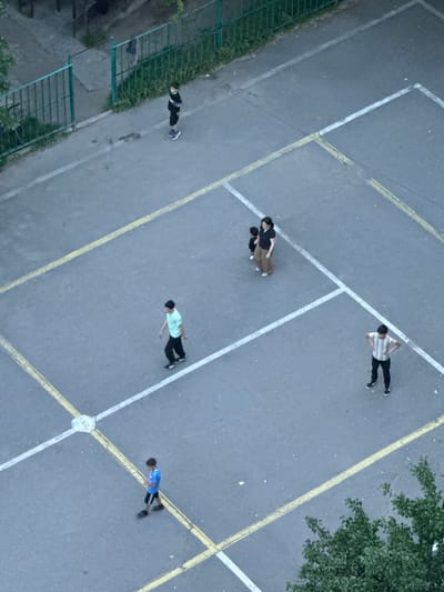 An aerial, high-angle shot captures a concrete playground with white and yellow painted lines in Abovyan, Armenia. Six individuals are visible on the playground. In the upper left, a person wearing a black t-shirt and shorts stands near a green metal fence. To the right, a woman in a black t-shirt and brown pants stands with a small child in a black outfit. Below them, a person in a light blue t-shirt and black pants walks to the left. To the right of the woman and child, a person in a striped t-shirt and black pants stands with hands on hips. In the lower left, a boy in a blue t-shirt and black shorts walks while looking down. The playground is surrounded by grass and some trees.