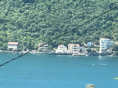 A wide shot shows a bay with calm, turquoise water in the foreground and a lush, green, tree-covered mountainside in the background. A line of buildings, predominantly white and beige with red-tiled roofs, is situated along the shoreline at the base of the mountains. These buildings appear to be a mix of residential homes and possibly small hotels or apartments. In the water, three small figures are visible on what appear to be paddleboards. The scene is likely Perast, Montenegro, given the distinctive mountainous backdrop and coastal architecture. A dark, thin cable is visible diagonally across the upper half of the image, obscuring parts of the mountainside and buildings. The lighting suggests a bright, sunny day.