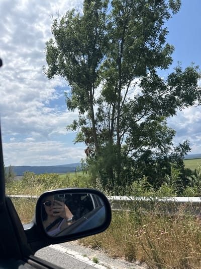 The image depicts a view from inside a car, looking outwards and reflected in the side mirror. In the foreground, the side of the car is visible, along with the road and a metal guardrail. The side mirror shows a selfie of a person wearing sunglasses and a light purple top, holding a phone. Behind the car, the landscape features a large tree with green foliage, a field, and a hilly horizon under a cloudy blue sky. The area appears to be rural, possibly in or near the city of Byala, Bulgaria, given the vegetation and terrain.