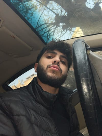 A young man with dark hair and a full beard is sitting inside a car. He wears a black puffer jacket. A black steering wheel is visible in the foreground. Through the car's windshield and side window, trees and buildings can be seen. The sun visor above is visible, with some wear and tear. The photo is taken at an unusual angle, looking slightly upward at the man.