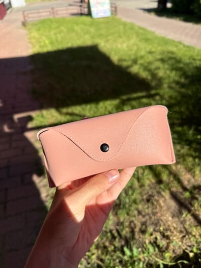 The image shows a close-up of a person's hand holding a light pink, hard-shell sunglasses case. The case has a snap closure with a black button. The background is blurred and appears to be an outdoor setting with green grass, shadows suggesting sunlight filtering through leaves, and a glimpse of a brick pathway. The lighting indicates it is daytime, likely sunny given the strong shadows. The overall impression is casual and relaxed, with the focus solely on the object being held. There is no visible text in the image.