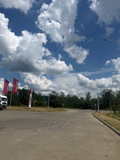 This image captures a roadside scene with a bright blue sky filled with dramatic white and gray clouds. The perspective is from a slightly elevated position, looking down onto a paved area that appears to be a parking lot or a roadside service area.
The primary subjects include a white truck partially visible in the foreground, and several red flags planted on poles. One flag, further away and higher up, displays white text that appears to be a logo or brand name, possibly Cyrillic. The closest flags also have red banners with white text that is partially obscured by the angle and the flag's movement. Behind these, a tall, vertical sign with red and white sections is visible, featuring multiple displays or indicators, suggesting it could be a price board or a service indicator, possibly related to fuel prices given the truck's presence.
The scene is set against a backdrop of dense green trees and foliage. A paved road or parking lot occupies the right side of the image, with white markings indicating parking spaces. The ground to the left of the parking lot is covered in dry, yellowish grass, with more trees beyond.
In the distance, along the roadside, a traffic signal or directional sign is visible, featuring a blue square with a white arrow pointing left, and a red circular sign. A street lamp with a metallic fixture is also present.
The lighting suggests it is daytime, likely late morning or afternoon, with the sun casting shadows and illuminating the clouds. The weather appears to be partly cloudy and warm, given the dry grass and the presence of the clouds. The overall impression is that of a functional, outdoor service or rest area along a road in a rural or semi-rural setting in Apsheronsk, Russia, given the provided location context. No specific activity or event is depicted beyond the static elements of the scene.