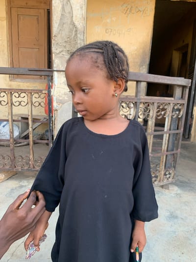 A young, dark-skinned child with dark braided hair, styled in cornrows across the top and loose braids on the sides, stands in an outdoor setting in Okene, Nigeria. The child wears a black, long-sleeved tunic with a round neckline and small green hoop earrings, gazing downwards and to the right. An adult's darker-skinned hand is visible, adjusting the left sleeve of the child's tunic. The child's right hand holds a multicolored pencil or crayon, while the left hand appears to hold a small, folded piece of patterned material. The background consists of a light brown, weathered building wall with a textured, possibly stucco finish. Faded black text and numbers, including "070449...", are visible written on the wall. A light brown wooden door is partially visible on the left side of the frame. Across the mid-ground, a dark, ornate metal railing with scrolled patterns is present, behind which some wooden debris or planks are visible. The ground beneath is a light-colored, flat surface.