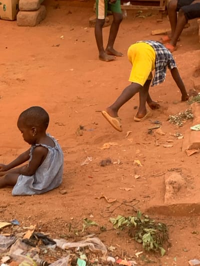 Three children are on reddish-brown soil. One child wears a light blue dress and bends forward, looking down. Another child wears yellow shorts and a blue-and-white checkered shirt, lying on their stomach. A third child is partly visible with green shorts. Scattered debris is nearby.