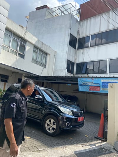 The image shows a man in a black shirt standing in front of a black SUV parked in a paved driveway. The man appears to be talking to someone inside the SUV, who is visible but not fully identifiable. The SUV is parked in front of a white building with large windows. There is a blue banner with Indonesian writing on the side of the building. The scene appears to be located in a residential or commercial area in Cimahi, Indonesia. The coordinates provided suggest that the location is in the neighborhood of Cimahi, West Java, near the intersection of Jalan Setiabudi and Jalan Jatihandap. The pavement of the driveway is made of cobblestone and the surroundings are clear. There is a small orange cone to the right of the image, partially obscured by a metal gate. The sky is partly cloudy.