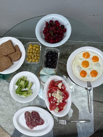 A top-down, eye-level, medium shot captures a glass-topped table laden with a breakfast spread. The scene is set outdoors, likely on a balcony or patio in Beşiktaş, Turkey, with a textured grey wall visible in the background.

Dominating the frame is a white plate of fried eggs, showcasing six distinct yolks surrounded by cooked whites. To its left, a white bowl overflows with ripe, dark red cherries. Adjacent to the cherries, a small glass container holds a cluster of green olives, and another holds black olives.

Below these, a white oval plate displays several slices of dark brown toast. Further down, a smaller white plate is filled with sliced cucumber sticks. To the right of the cucumber, a white oval dish contains sliced tomatoes, generously topped with crumbled white cheese. At the bottom of the frame, another white oval plate holds thin slices of red and white marbled salami.

Two silver forks are positioned on the table, one to the right of the fried eggs and another below the salami. The overall impression is that of a freshly prepared, healthy breakfast, ready to be enjoyed.