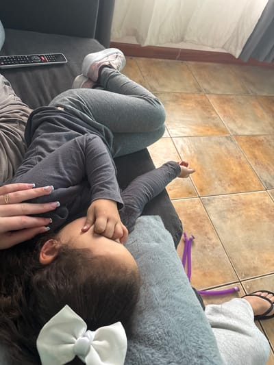 The image shows a close-up, overhead view of two people lying down on a couch, with their legs extending towards a tiled floor. The person in the foreground, a child, is lying on their side, with their head resting on a gray blanket. Their eyes are closed, and their hand is covering their face. A white bow is visible in their dark hair. To the left, an adult's hand is resting on the child's head, adorned with a ring and manicured nails. The adult is wearing gray sweatpants and black flip-flops. In the background, a remote control rests on the couch, and sheer white curtains hang near a window. The tiled floor is a warm, earthy tone. The scene appears to be an indoor domestic setting. There is no information in the image to suggest the location is in Iquique, Chile.