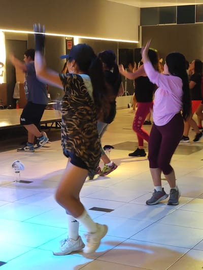 The image depicts an indoor group fitness class, likely a dance or Zumba session, taking place in Tacna, Peru. Several people, predominantly women, are actively participating, performing energetic movements with raised arms.
In the foreground, a woman with a black baseball cap and long dark hair, seen from behind, wears a distinctive tiger-print t-shirt, black shorts, white socks, and white sneakers. Her arms are raised high, showing a moment of dynamic action. To her left, in the mid-ground, a man in a blue t-shirt, black shorts, and blue and white Nike Air Jordan high-top sneakers also has his arms lifted, mirroring the energetic pose. Further to the right, another woman in a light pink long-sleeved top, dark red capri leggings, and gray and pink sneakers is also engaged in the exercise, with the Adidas logo visible on her white socks. Other participants are visible in the background, all actively moving.
The setting is a modern fitness studio or gym with a light-colored tiled floor, illuminated by vibrant blue and cyan lights, creating an energetic and dynamic atmosphere. Overhead lighting also contributes to the bright environment. In the background, black speakers on stands and some benches or platforms can be seen. Water bottles are placed on the floor, one clearly visible in the foreground with a blue cap and a light-colored item on top, suggesting hydration breaks during the intense activity. The participants appear focused and engaged, indicative of an enjoyable and energetic workout session.