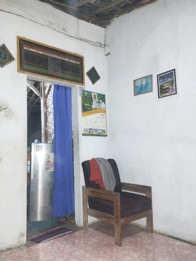 This image captures a static interior view of a modest room, likely a living area or entry hall, within a home in Kecamatan Arjasa, Indonesia. The walls are painted white, showing some signs of wear and minor cracking near the ceiling. The floor is covered with reddish-brown patterned tiles. Above, exposed wooden beams and rafters are visible, characteristic of traditional Indonesian architecture.
On the left side of the room, a doorway is partially obscured by a vibrant blue curtain, revealing a silver-colored refrigerator and a glimpse into an adjacent space. A small, striped purple and brown rug lies on the floor in front of the doorway.
The walls are decorated with various items. Centered above the doorway is a large, rectangular framed artwork featuring intricate Arabic calligraphy, with the word "Allah" prominently displayed in the middle. To its left and right are two smaller, square-framed decorative pieces with circular Islamic patterns. On the right wall, two framed pictures are hung: one showing a figure against a light blue background, and the other depicting a building with a green roof, possibly a mosque or shrine. Below these pictures hangs a wall calendar for the year "2024," featuring an image of several men in white Islamic attire.
A simple wooden armchair with dark upholstery is placed against the right wall, with red and grey clothes casually draped over its back. An electrical outlet or light switch is visible on the left wall.
The lighting suggests it is evening or night, with artificial illumination casting a soft glow within the room, and the area visible through the doorway appears dark. No people are present, and no specific activity is taking place, creating a calm and ordinary domestic scene. The overall impression is a lived-in, unpretentious home setting.