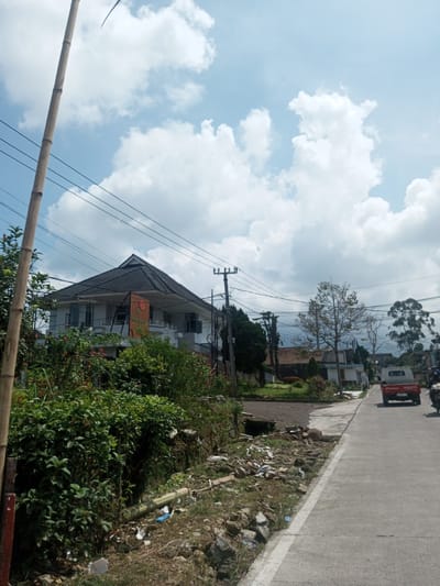 The image shows a street scene in a residential area. A two-story house with a gray roof is visible behind dense green vegetation. Overhead power lines stretch across the sky, which is filled with large, puffy white clouds. Debris and vegetation line the edge of the road, which is made of gray asphalt. A red pickup truck drives down the street, and a motorbike follows behind. A bamboo pole stands tall on the left side of the frame.