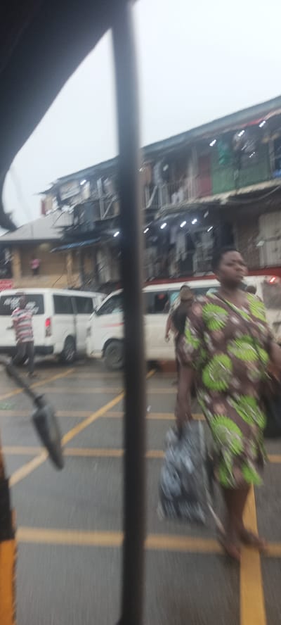 The scene is a street in Umuahia, Nigeria, appearing to be a market area with buildings in the background. The street has yellow lane markings. There are white vans parked along the side of the road. A woman in a green and brown patterned dress is walking barefoot across the street, carrying a large bag. The view is obscured by what seems to be a pole or part of the vehicle from which the photograph was taken. A man in a striped shirt is walking near one of the vans.