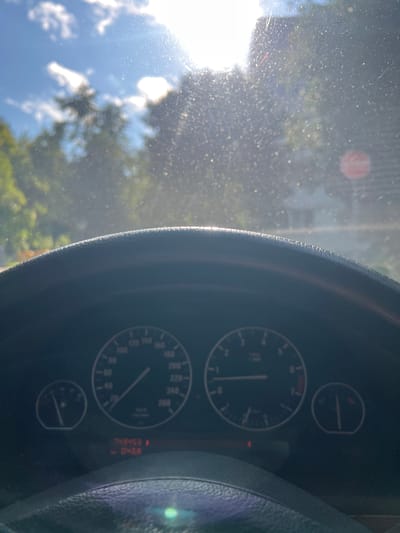 The image shows a view from inside a car, looking out through the windshield. The sun is bright and high in the sky, with some clouds present. Trees and buildings are visible in the background, but they are out of focus due to the glare and the view through the windshield. The steering wheel and dashboard are in the foreground, with the speedometer and tachometer visible. The car's odometer displays "243453" kilometers, and the trip meter shows "0438". The image appears to have been taken in a rural or suburban area.