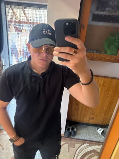 This is a portrait-oriented, low-angle, full-body selfie taken indoors. The subject is a young person wearing a black baseball cap, gold-rimmed sunglasses, a black t-shirt, and black ripped jeans. They are wearing a beaded bracelet on their left wrist and a ring on their right ring finger. They are holding a black smartphone in their left hand to take the picture. The background shows a wooden floor and a wooden paneled wall, with a glimpse of a rug and some footwear, including a white flip-flop and dark sneakers, suggesting a domestic setting. The lighting appears to be natural, possibly from a window, and the overall mood is casual and relaxed. The image is oriented vertically and appears to have been taken on a sunny day, as suggested by the brightness and the reflection in the sunglasses. The text on the baseball cap is in a stylized script, but is not clearly legible. The location context is Kostinbrod, Bulgaria.