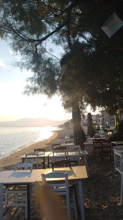 A beachside cafe in Neoi Epivates, Greece, is captured at sunset. The scene is set on a sandy beach with the calm sea in the background. Several tables and chairs, some white and some dark wood, are arranged on the sand, suggesting a dining area. Two large, closed umbrellas are visible, casting shadows. Buildings line the distant shore, and a mountain range can be seen in the hazy distance. The sun, low in the sky, casts a warm glow and creates reflections on the water. The leaves and branches of a large tree overhang the scene from the top left. The lighting suggests it is late afternoon or early evening. There are no people clearly visible in the image.