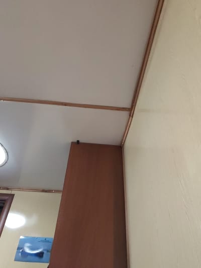 The image shows an interior view looking upwards towards a ceiling and a corner where two walls meet. The ceiling is a light color, possibly white or off-white, and appears to be a smooth surface. Along the edge where the ceiling meets the walls, there is a thin wooden trim, likely for finishing. The walls are also a light, neutral color, with one wall exhibiting a wood-grain texture. A tall, dark brown wooden structure, possibly a cabinet or part of a room divider, occupies a significant portion of the right side of the frame, extending upwards.
On the left side of the image, a circular light fixture with a white frame is visible, suggesting an internal light source. Below this, on the wall, a piece of paper or poster is attached. It appears to be a photograph or print of a large boat or ship on water, with some text in blue at the top. The overall lighting suggests it is daytime, but the interior setting makes it difficult to determine the exact time of day or weather conditions outside. There are no people visible in the image. The setting appears to be a room or hallway, possibly in a residential or commercial building. The image is taken from a low angle, emphasizing the ceiling and architectural elements.