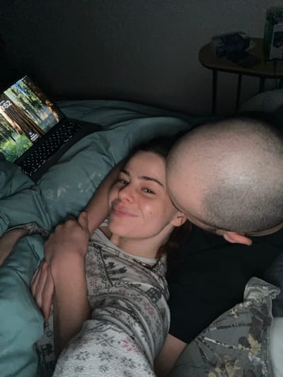 A cozy, intimate scene depicts a couple in bed. A woman with a gentle smile rests against blue pillows, wearing a patterned pajama top. She is embraced by a man who is kissing her head. A laptop sits open nearby, displaying a forest scene. The overall lighting suggests a dim, relaxed atmosphere.
