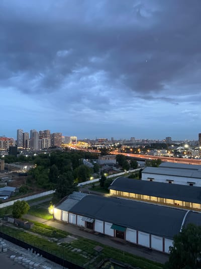 This is an aerial, low-light photograph of an industrial and residential area at dusk in Chelyuskinsky, Russia. In the foreground, there are two long, low-rise industrial buildings with dark, arched roofs and white walls. The building on the right has its front facing the viewer and shows a row of illuminated doorways. The building on the left, closer to the viewer, has a dark roof and white walls with red trim, and several garage-like doors. A single, bright light illuminates the area in front of this building. Beyond these structures, there is a dense area of green trees, and further in the distance, a highway overpass is visible, with its traffic lights creating streaks of orange light. The background is dominated by a cluster of tall, modern residential buildings, illuminated by interior lights. The sky is overcast with dark, heavy clouds, and a faint crescent moon is visible. The overall impression is one of an urban landscape transitioning into evening, with a blend of industrial and residential zones.