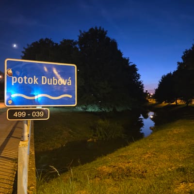 At night, a blue road sign with yellow lettering reads "potok Dubová" above a wavy line symbol and a smaller black sign with the numbers "499 - 039". The sign is attached to a concrete post. To the right of the sign, a narrow body of dark water reflects lights from the surrounding area, bordered by grassy banks and trees. The sky is a deep blue, with a sliver of moon visible in the upper left corner. The location is identified as Piešťany, Slovakia.
