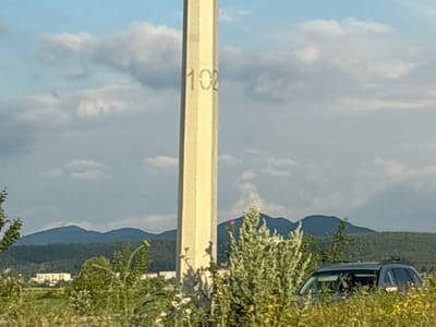 The image depicts an outdoor scene with a tall, light-colored concrete pillar in the foreground. The number "102" is visible, stenciled on the pillar. The pillar is positioned in a grassy area with various plants and bushes, some of which have small flowers.
In the background, a dark-colored SUV is partially visible, peeking through the foliage. Beyond the immediate foreground, there is a dense line of trees and then a series of rolling mountains under a cloudy sky. The sky is a mix of blue and gray clouds, suggesting a partly cloudy day. On the left side of the background, several buildings are visible, indicating a nearby settlement.
The overall setting appears to be rural or semi-rural, possibly a roadside or an open field. The lighting suggests it is daytime, possibly late afternoon or early evening, given the angle of the light and the soft colors. There are no people visible in the image. The scene is peaceful and natural. The presence of the numbered pillar might suggest a boundary marker, mile marker, or some form of survey point. The location context is Akhalsopeli, Georgia.