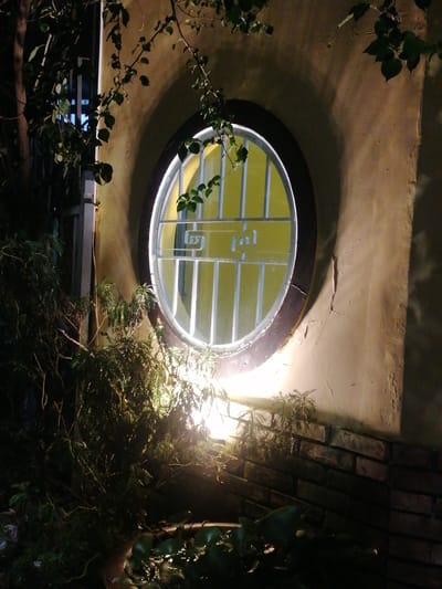 This is a nighttime photograph taken at Xuân Hương Lake in Đà Lạt, Vietnam. The scene features a rustic, circular window set into a textured, cream-colored wall. The window is illuminated from within by a bright yellow light, and a decorative white metal grate with a spiral motif covers the glass. Lush, dark green foliage, including tree branches with leaves and smaller bushy plants, partially obscures the view of the wall and window, casting shadows and creating a dramatic, atmospheric effect. The lower portion of the wall is constructed of dark brick. The lighting suggests an outdoor setting, likely a garden or courtyard adjacent to a building.