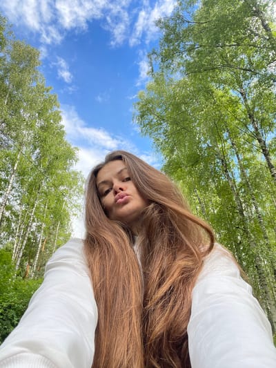A young woman with long, brown hair is captured in an upside-down selfie taken outdoors. She is wearing a white long-sleeved shirt. Her face is turned towards the camera, with her lips slightly pursed. The background consists of a vibrant blue sky with scattered white clouds, and the lush green canopy of trees, likely birch trees given the location context of Заречный, Russia. The sunlight suggests it is daytime, possibly late morning or afternoon, with bright but not harsh lighting. The overall mood appears casual and relaxed, with the woman enjoying a moment outdoors amongst nature. No visible text is present in the image.